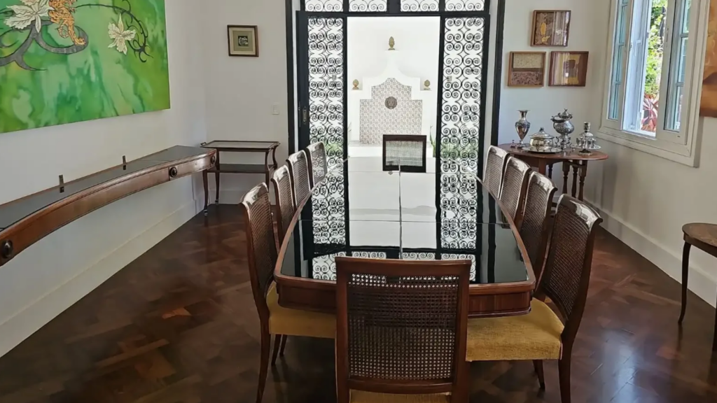 Sala de jantar com diversos quadros nas paredes e uma mesa longa de vidro e madeira ao centro com cadeiras estofadas e encosto de tela.