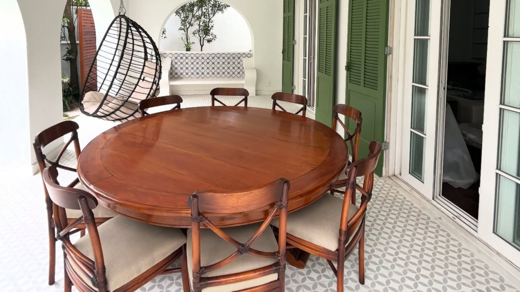 Espaço externo com mesa redonda de madeira robusta e cadeiras de encosto fino em x e cadeira arredondada suspensa.