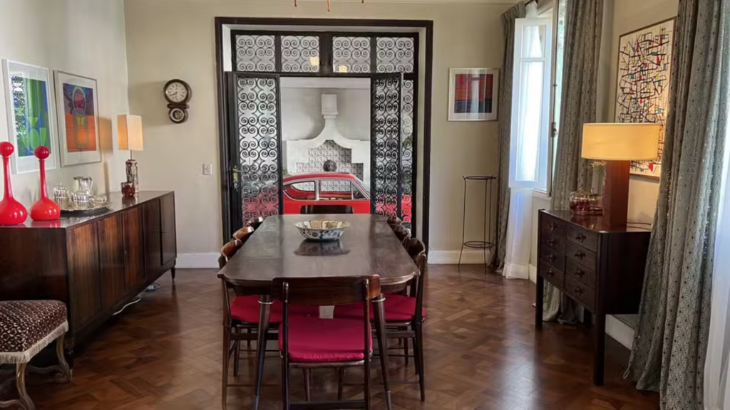 Sala de jantar com porta de vidro e arabescos de metal, decorações de cores quentes, mesa robusta de madeira maciça e cadeiras finas de estofado vermelho.