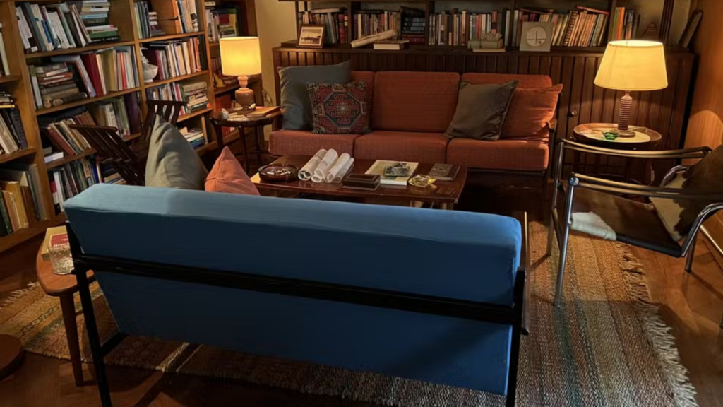 Escritório simples com livros e sofás, um laranja e o outro azul, de base metálica fina e uma cadeira de base igual, mas acento e encosto retos de couro.