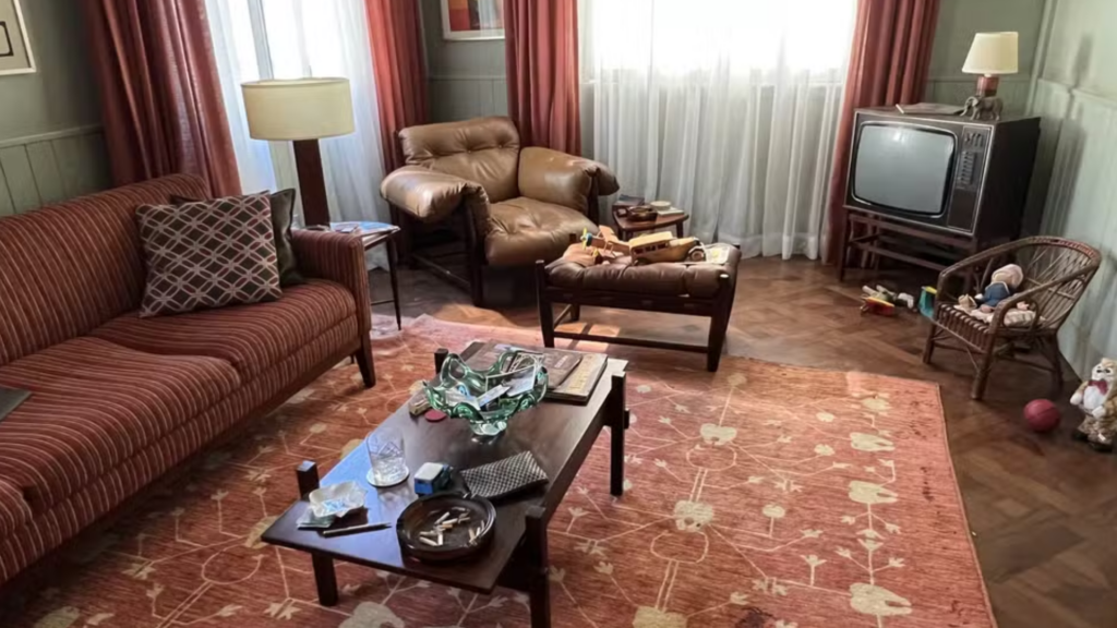 Sala de estar com objetos espalhados no chão, cadeiras e mesa, sofá avermelhado e poltrona de couro bege aos cantos.