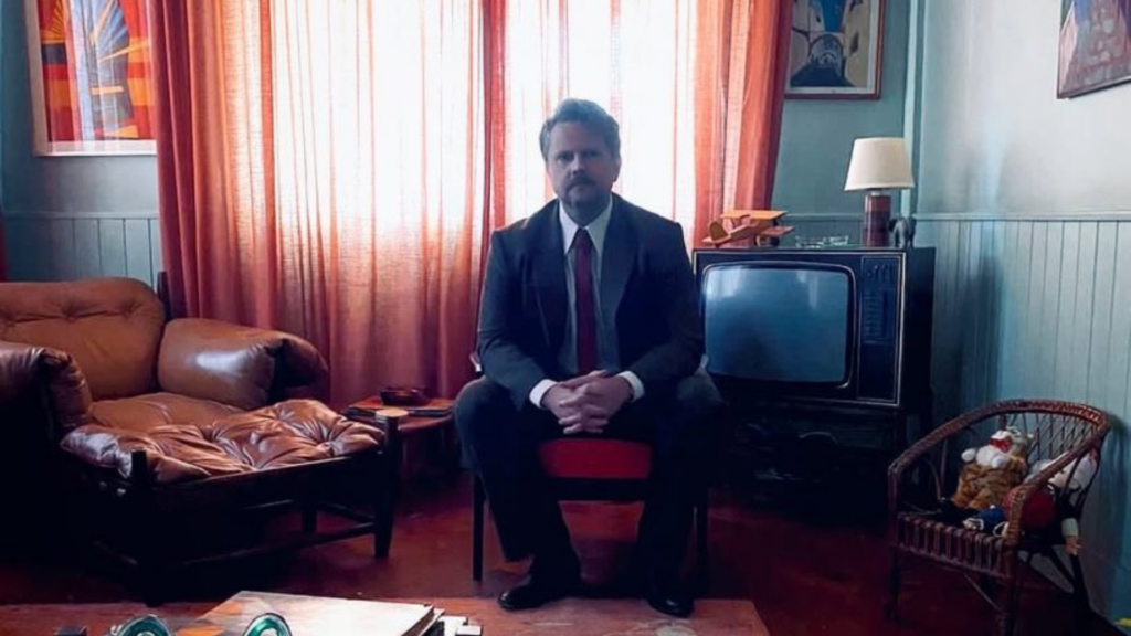 Personagem Marcelo Rubens Paiva, interpretado por Selton Mello, sentado ao centro de uma sala de cortina laranja.
