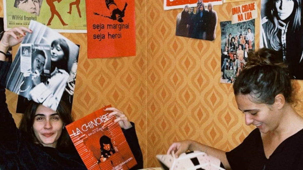 Quarto com foco na parede de papel de parede amarelo em detalhes em laranja com diversos pôsteres e colagens.