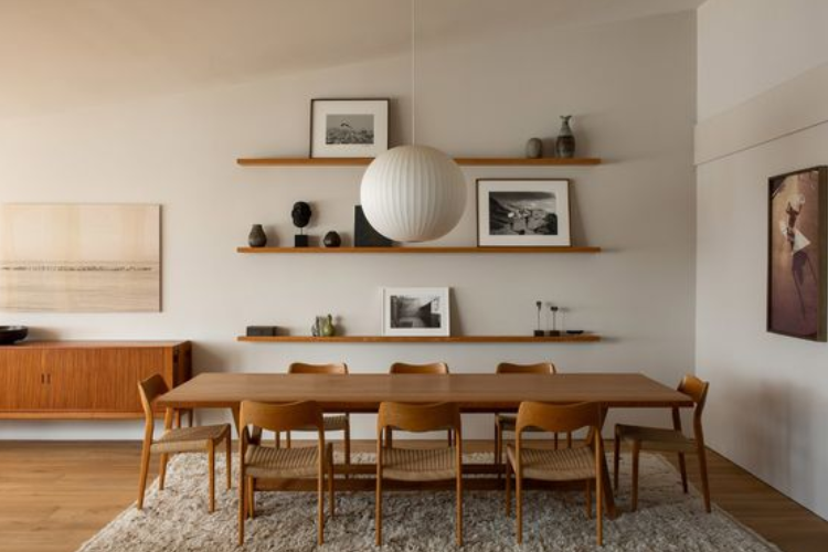 Grande sala de jantar com uma extensa mesa retangular e 8 cadeiras, ambas de madeira, com decoração simples de quadros e enfeites de prateleira